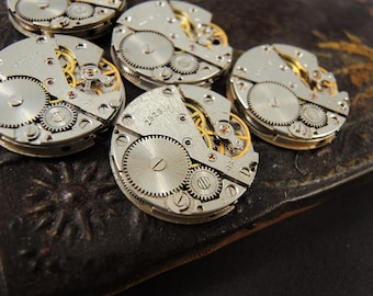 May include: Close-up of several disassembled watch movements. The silver-toned mechanisms feature intricate gears, tiny screws, and golden-colored components. The number "2528" is visible on one of the movements. The background is a dark, textured surface.
