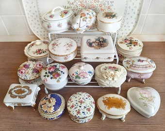 May include: A collection of various porcelain trinket boxes in round, square, and heart shapes. The boxes feature floral patterns, gold trim, and decorative lids. Some boxes have small feet, while others are plain. They are arranged on a white rack.