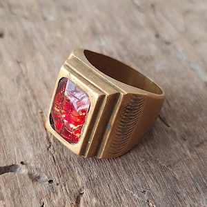 May include: A vintage brass ring with a rectangular, red gemstone. The ring has a square face with a raised bezel setting the stone. The band is thick and features etched detailing on the side. The ring is set on a wooden surface.