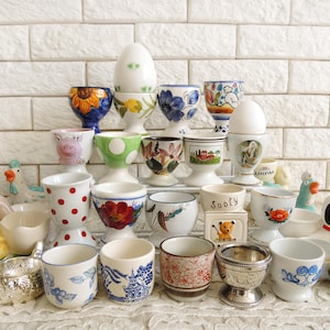 Peut inclure: Une collection de coquetiers décoratifs de différents styles, notamment floraux, à pois et animaliers. Certains coquetiers contiennent des œufs. Les coquetiers sont en céramique et en porcelaine, avec une variété de couleurs et de motifs.