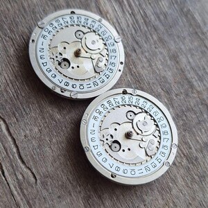 K&ouml;nnte beinhalten: Zwei Vintage-Uhrwerkkomponenten mit sichtbaren Zahnr&auml;dern und Datumsanzeigen. Die silberfarbenen Metallteile weisen einen wei&szlig;en Ring mit schwarzen Zahlen von 1 bis 31 auf, die die Tage des Monats angeben. Der Hintergrund ist eine Holzoberfl&auml;che.