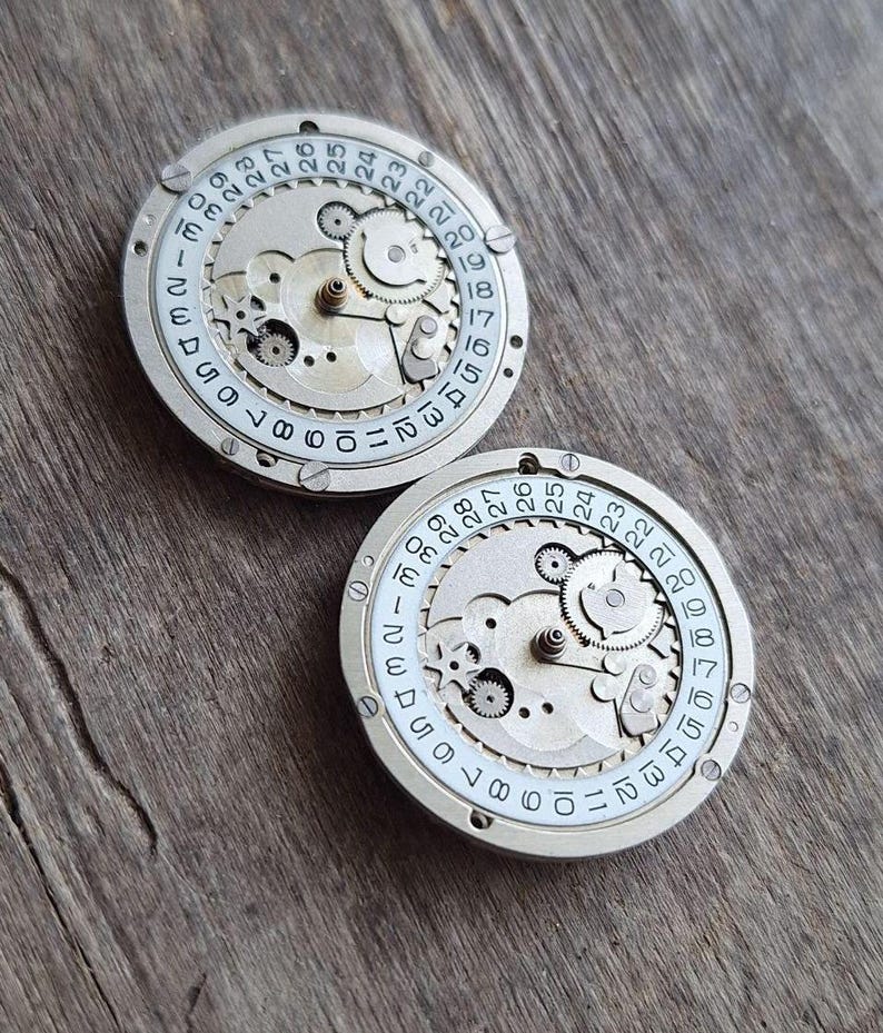 K&ouml;nnte beinhalten: Zwei Vintage-Uhrwerkkomponenten mit sichtbaren Zahnr&auml;dern und Datumsanzeigen. Die silberfarbenen Metallteile weisen einen wei&szlig;en Ring mit schwarzen Zahlen von 1 bis 31 auf, die die Tage des Monats angeben. Der Hintergrund ist eine Holzoberfl&auml;che.