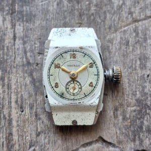 Peut inclure: Montre-bracelet Zvezda vintage avec un bo&icirc;tier rectangulaire argent&eacute;. Le cadran est blanc cass&eacute; avec des aiguilles et des chiffres dor&eacute;s. La montre a un petit cadran de secondes et une couronne textur&eacute;e. Pos&eacute;e sur une surface en bois vieilli.