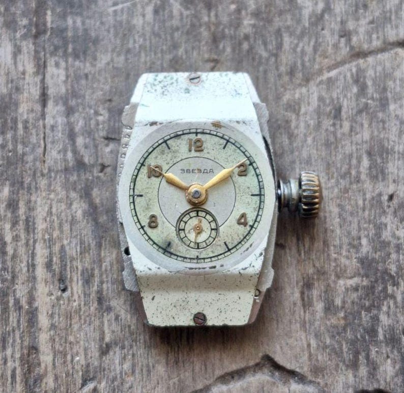 Peut inclure: Montre-bracelet Zvezda vintage avec un bo&icirc;tier rectangulaire argent&eacute;. Le cadran est blanc cass&eacute; avec des aiguilles et des chiffres dor&eacute;s. La montre a un petit cadran de secondes et une couronne textur&eacute;e. Pos&eacute;e sur une surface en bois vieilli.