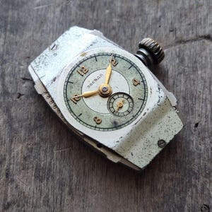 Peut inclure: Montre-bracelet vintage avec un bo&icirc;tier argent&eacute; vieilli et un cadran vert d&eacute;lav&eacute;. Le cadran pr&eacute;sente des aiguilles et des chiffres dor&eacute;s, avec un petit cadran secondaire. La montre est partiellement d&eacute;mont&eacute;e, montrant des signes d'&acirc;ge.