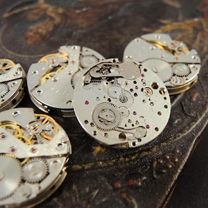 May include: Close-up of several disassembled watch movements, showcasing intricate gears, springs, and jewels. The silver-toned components are arranged on a dark, textured surface, highlighting the precision of the watchmaking craft.