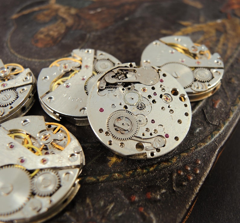 May include: Close-up of several disassembled watch movements, showcasing intricate gears, springs, and jewels. The silver-toned components are arranged on a dark, textured surface, highlighting the precision of the watchmaking craft.