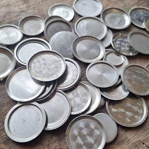 May include: A collection of various sized, circular, silver-toned watch case backs. The backs are made of metal and have a brushed or textured finish. They are scattered on a wooden surface.