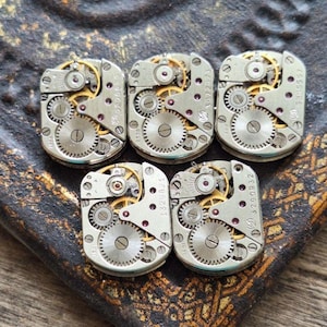 May include: Five vintage watch movements with intricate gears and silver-toned metal components. The watch parts are arranged on a textured, dark surface, showcasing the detailed mechanics of the timepieces. The gears are gold-toned.
