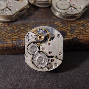 May include: Close-up of several vintage watch movements. The central movement is silver-toned with visible gears, jewels, and a balance wheel. The background shows a dark, textured surface and other watch components.