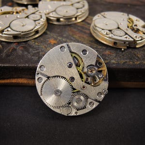 May include: Close-up of several vintage watch movements, showcasing intricate gears, screws, and other mechanical components. The movements are silver and gold-toned, with a focus on the detailed inner workings of the timepieces. The image highlights the craftsmanship of antique watchmaking.