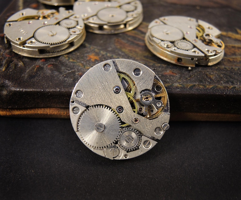 May include: Close-up of several vintage watch movements, showcasing intricate gears, screws, and other mechanical components. The movements are silver and gold-toned, with a focus on the detailed inner workings of the timepieces. The image highlights the craftsmanship of antique watchmaking.