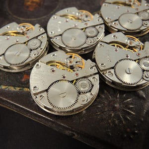 May include: A collection of vintage watch movements, showcasing intricate gears, springs, and other mechanical components. The silver-toned metal contrasts with the gold-colored gears and red jewels. These watch parts are arranged on a dark, textured surface.