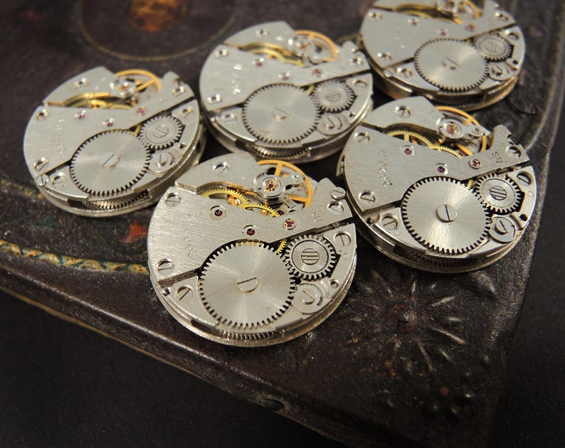 May include: A collection of vintage watch movements, showcasing intricate gears, springs, and other mechanical components. The silver-toned metal contrasts with the gold-colored gears and red jewels. These watch parts are arranged on a dark, textured surface.