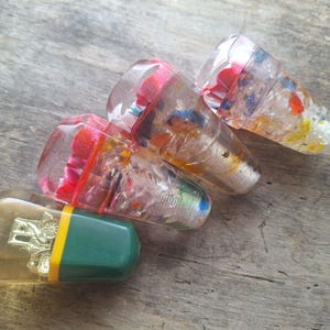 May include: A collection of decorative bottle stoppers. The stoppers are made of clear resin, some with colorful inclusions of red, blue, and yellow. One stopper has a green and yellow cap with a gold emblem.