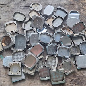 Puede incluir: Una colección de traseras de relojes antiguos de diversas formas y tamaños, incluyendo rectangulares, cuadrados y ovalados. Los componentes metálicos de los relojes tienen un aspecto desgastado y envejecido, con algunos mostrando signos de oxidación y desgaste. Las traseras de los relojes están esparcidas sobre una superficie de madera.