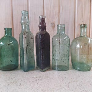 May include: Five antique glass bottles of varying shapes and colors. The bottles range from green to clear and purple, with some showing signs of age and wear. The bottles are arranged in a row against a light wood background.