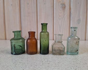 May include: Five antique glass bottles in different shapes and colours. The bottles vary from green to amber and clear, with visible imperfections. The bottles are arranged in a row, showcasing their unique designs and aged appearance.