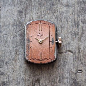 Puede incluir: Reloj de pulsera rectangular vintage con esfera color cobre, manecillas e indicadores horarios dorados. El reloj tiene una caja plateada y una corona dorada. La esfera muestra la palabra cir&iacute;lica "Луч" y el texto "Сделано en la URSS".