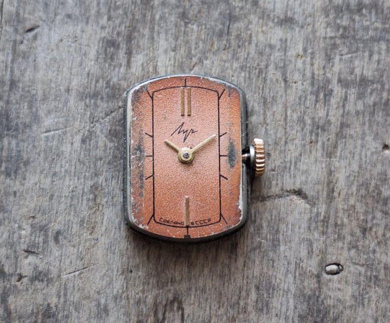 Puede incluir: Reloj de pulsera rectangular vintage con esfera color cobre, manecillas e indicadores horarios dorados. El reloj tiene una caja plateada y una corona dorada. La esfera muestra la palabra cir&iacute;lica "Луч" y el texto "Сделано en la URSS".