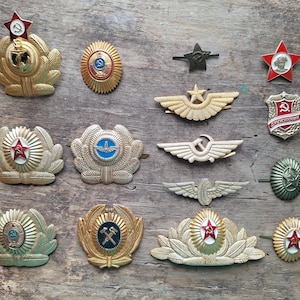 Puede incluir: Una colección de insignias y distintivos militares antiguos, con colores dorados, rojos y verdes. El surtido incluye estrellas, alas y emblemas con símbolos de martillo y hoz, y una estrella roja.