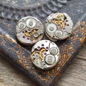 May include: Three vintage watch movements with visible gears and intricate mechanical components. The silver-toned metal pieces feature gold-colored gears and small red jewels. The watch parts are arranged on a dark, textured surface.