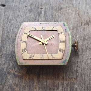 Peut inclure: Cadran de montre vintage de forme rectangulaire avec un cadre dor&eacute;. Le cadran affiche des chiffres romains et des aiguilles dor&eacute;es. Le cadran de la montre est un m&eacute;lange de bronze, d'or et de vert. La couronne de la montre est sur le c&ocirc;t&eacute; droit.