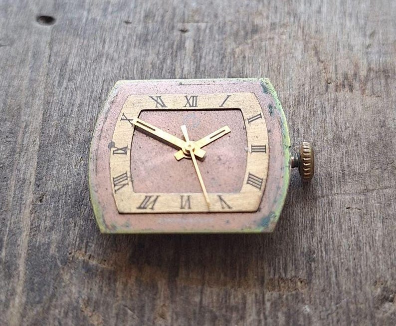 Peut inclure: Cadran de montre vintage de forme rectangulaire avec un cadre dor&eacute;. Le cadran affiche des chiffres romains et des aiguilles dor&eacute;es. Le cadran de la montre est un m&eacute;lange de bronze, d'or et de vert. La couronne de la montre est sur le c&ocirc;t&eacute; droit.