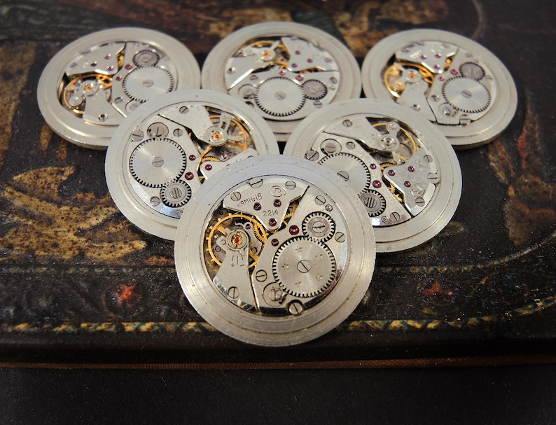 K&ouml;nnte beinhalten: Sechs Vintage-Uhrwerke mit freiliegenden Zahnr&auml;dern, Federn und Juwelen. Die silberfarbenen Metallkomponenten sind kreisf&ouml;rmig angeordnet und zeigen die komplizierten Innenarbeiten der Zeitmesser. Die Zahnr&auml;der sind goldfarben und die Juwelen sind rot.