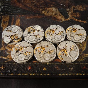 Peut inclure: Sept mouvements de montre vintage avec des engrenages complexes, du m&eacute;tal argent&eacute; et des accents dor&eacute;s. Les pi&egrave;ces de la montre sont dispos&eacute;es sur une surface sombre et &agrave; motifs, mettant en valeur la m&eacute;canique d&eacute;taill&eacute;e des garde-temps. Une collection de composants horlogers.