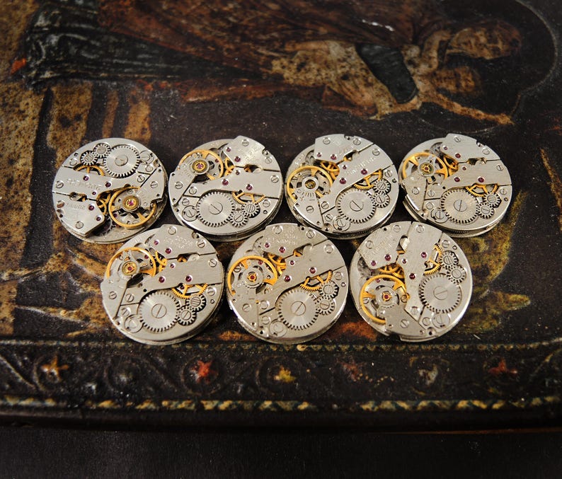 Peut inclure: Sept mouvements de montre vintage avec des engrenages complexes, du m&eacute;tal argent&eacute; et des accents dor&eacute;s. Les pi&egrave;ces de la montre sont dispos&eacute;es sur une surface sombre et &agrave; motifs, mettant en valeur la m&eacute;canique d&eacute;taill&eacute;e des garde-temps. Une collection de composants horlogers.
