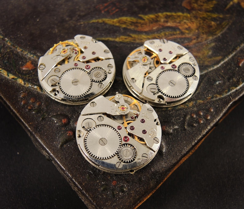 Pode incluir: Tr&ecirc;s movimentos de rel&oacute;gio vintage circulares, com engrenagens intrincadas, componentes de metal prateado e pequenas joias vermelhas. As pe&ccedil;as do rel&oacute;gio s&atilde;o exibidas em uma superf&iacute;cie escura e texturizada.