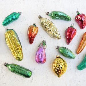 May include: A collection of vintage glass vegetable ornaments in various colors. The ornaments include a carrot, corn, peppers, cucumber, and eggplant. Each ornament has a metal cap for hanging. The ornaments are arranged on a white surface.