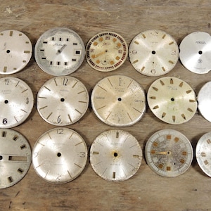 Puede incluir: Una colección de esferas de reloj vintage de varios tamaños y diseños. Las esferas son principalmente de color plateado con números y marcas negros. Algunas muestran signos de edad y desgaste, con pátina y decoloración visibles. La colección está dispuesta sobre una superficie de madera.
