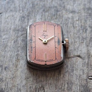 Puede incluir: Reloj de pulsera rectangular vintage con esfera de color cobre, manecillas doradas y corona texturizada. La esfera del reloj tiene l&iacute;neas verticales y texto cir&iacute;lico. El reloj est&aacute; sobre una superficie de madera.