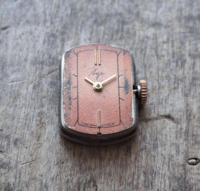 Puede incluir: Reloj de pulsera rectangular vintage con esfera de color cobre, manecillas doradas y corona texturizada. La esfera del reloj tiene l&iacute;neas verticales y texto cir&iacute;lico. El reloj est&aacute; sobre una superficie de madera.
