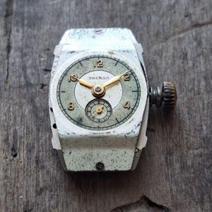 Peut inclure: Montre-bracelet Zvezda vintage avec un bo&icirc;tier rectangulaire blanc et un cadran vert d&eacute;lav&eacute;. La montre pr&eacute;sente des aiguilles et des chiffres dor&eacute;s, avec un petit cadran de secondes. La montre est pos&eacute;e sur une surface en bois vieilli.