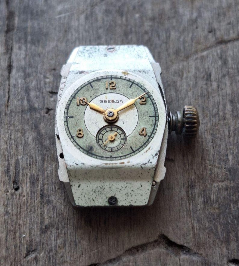 Peut inclure: Montre-bracelet Zvezda vintage avec un bo&icirc;tier rectangulaire blanc et un cadran vert d&eacute;lav&eacute;. La montre pr&eacute;sente des aiguilles et des chiffres dor&eacute;s, avec un petit cadran de secondes. La montre est pos&eacute;e sur une surface en bois vieilli.