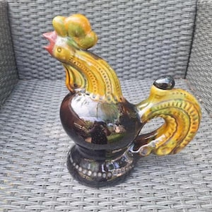 Puede incluir: Jarra de cerámica con forma de gallo y acabado brillante. La jarra presenta un cuerpo marrón, cuello amarillo y verde y cresta roja. El asa y el pico están integrados en el diseño del gallo. Es un artículo decorativo.