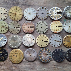 Pode incluir: Uma coleção de vinte e três mostradores de relógio vintage circulares em vários estados de desgaste, com diversos acabamentos metálicos incluindo tons de ouro, prata, preto e cobre, com marcadores de hora, números e janelas de data variados.