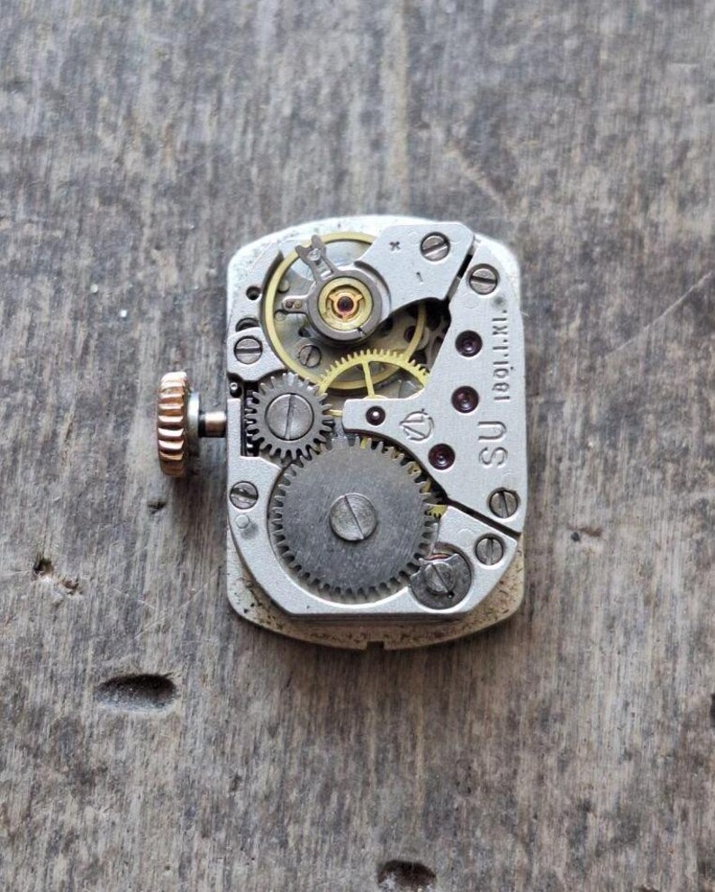 Puede incluir: Primer plano de un movimiento de reloj vintage. El mecanismo rectangular plateado presenta intrincados engranajes, tornillos y un volante dorado. Una peque&ntilde;a corona texturizada de oro rosa es visible en el lateral. El texto "SU 1901.1.XI." est&aacute; impreso en el mecanismo.