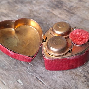 May include: A heart-shaped vanity case with a red textured exterior and a gold-toned interior. The case is open, revealing two small round gold-toned metal containers and a small circular brush with bright red bristles nestled inside the base.