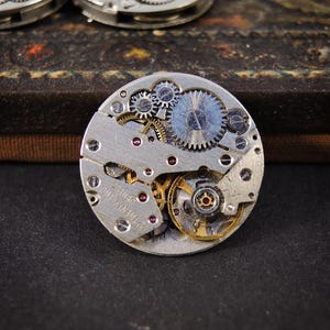 Könnte beinhalten: Nahaufnahme eines Vintage-Uhrwerks mit komplizierten Zahnrädern und Komponenten. Der silberfarbene Mechanismus weist eine komplexe Anordnung kleiner Zahnräder, Juwelen und anderer Teile auf. Der Text "22 TWENTY-TWO JEWELS" ist sichtbar.