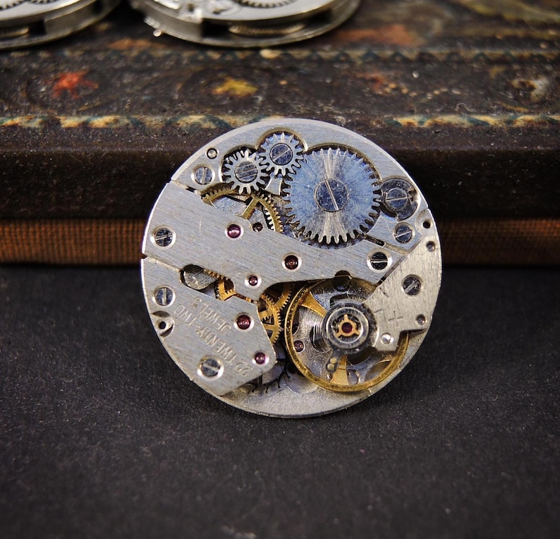 Peut inclure: Gros plan d'un mouvement de montre vintage avec des engrenages et des composants complexes. Le m&eacute;canisme argent&eacute; pr&eacute;sente un agencement complexe de petits engrenages, de bijoux et d'autres pi&egrave;ces. Le texte "22 TWENTY-TWO JEWELS" est visible.