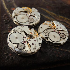 Pode incluir: Tr&ecirc;s movimentos de rel&oacute;gio vintage com engrenagens intrincadas, rodas e joias de rubi. Os mecanismos prateados est&atilde;o dispostos sobre uma superf&iacute;cie escura e texturizada, mostrando o artesanato detalhado das pe&ccedil;as do rel&oacute;gio.