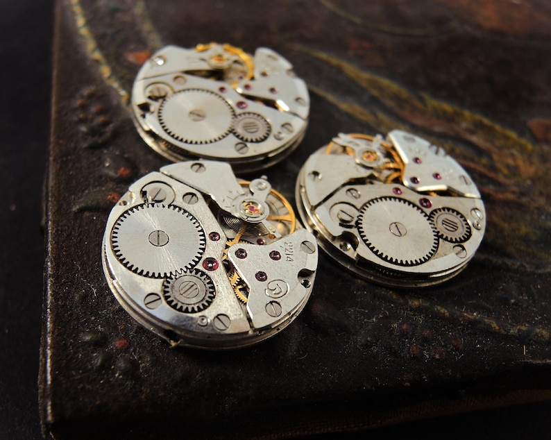 Pode incluir: Tr&ecirc;s movimentos de rel&oacute;gio vintage com engrenagens intrincadas, rodas e joias de rubi. Os mecanismos prateados est&atilde;o dispostos sobre uma superf&iacute;cie escura e texturizada, mostrando o artesanato detalhado das pe&ccedil;as do rel&oacute;gio.