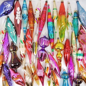 May include: A collection of assorted glass Christmas ornaments in various elongated, twisted, and teardrop shapes. The ornaments feature a vibrant palette of red, blue, green, gold, pink, and purple, with painted floral, striped, and glittered details.