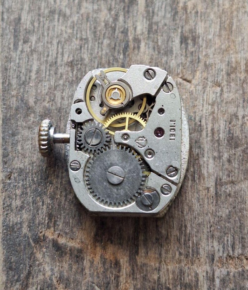 May include: Close-up of a vintage watch movement with intricate gears, a winding stem, and a small ruby jewel. The metal components are silver and gold-toned, set against a weathered wooden surface. The watch mechanism is oval-shaped.