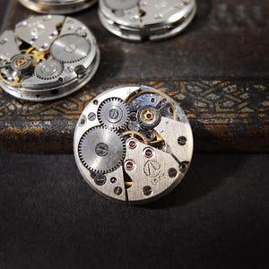 Könnte beinhalten: Nahaufnahme mehrerer zerlegter Uhrwerke, die komplizierte Zahnräder, Federn und andere mechanische Komponenten zeigen. Die Uhrteile sind silber- und goldfarben, mit Fokus auf die detaillten Innenarbeiten der Zeitmesser. Der Hintergrund ist dunkel.