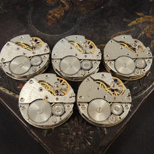 May include: Five vintage watch movements with visible gears and intricate mechanical details. The silver-toned components include small screws, wheels, and a balance wheel, with gold and red accents. The watch parts are arranged on a dark, textured surface.